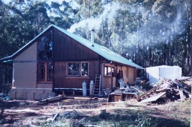 A rustic scene with smoke coming from the chimney of a mud brick and timber house surrounded by bush.