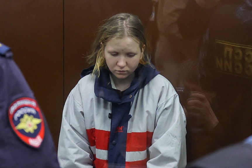 A woman in a white, blue and red jacket sits behind a glass wall in court.