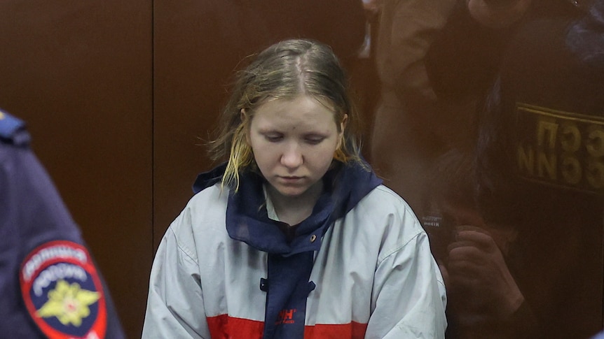 A woman in a white, blue and red jacket sits behind a glass wall in court.