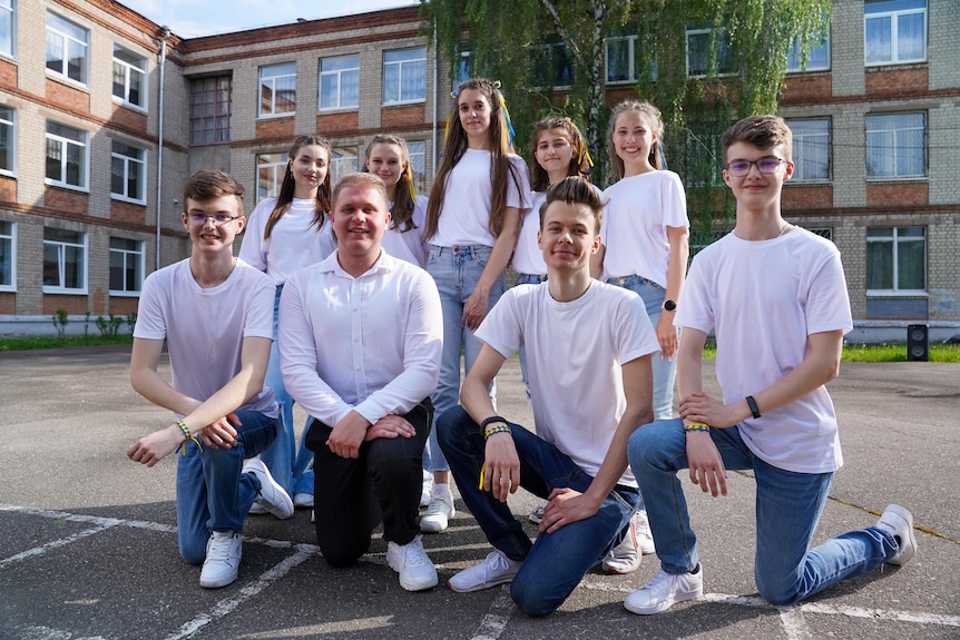 Nine teenagers wearing matching blue jeans and white tshirt and sneakers pose for a photo in front of their school building