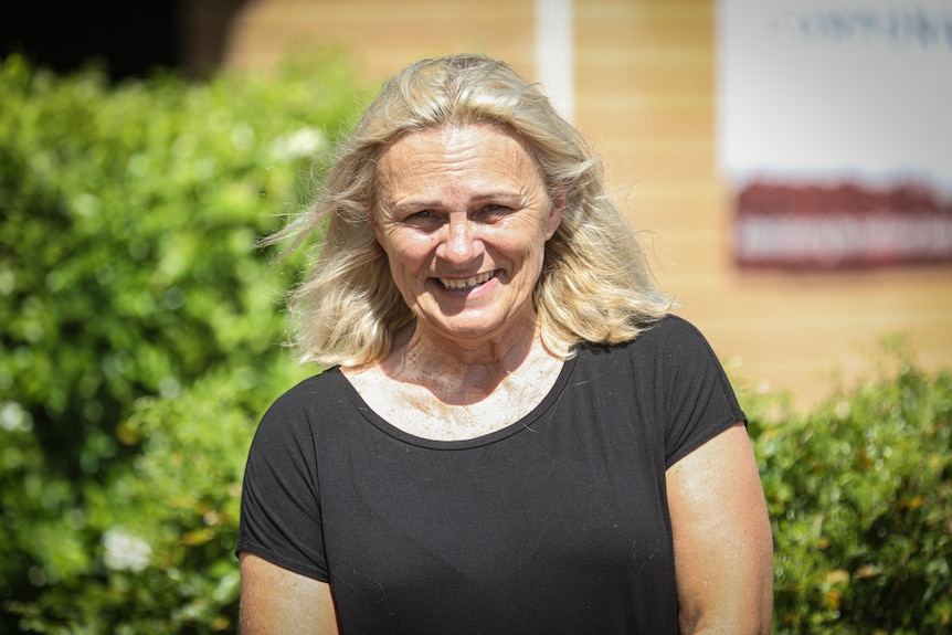 A middle-aged blonde woman smiles, wears a black T-shirt, blurred orange brick wall.