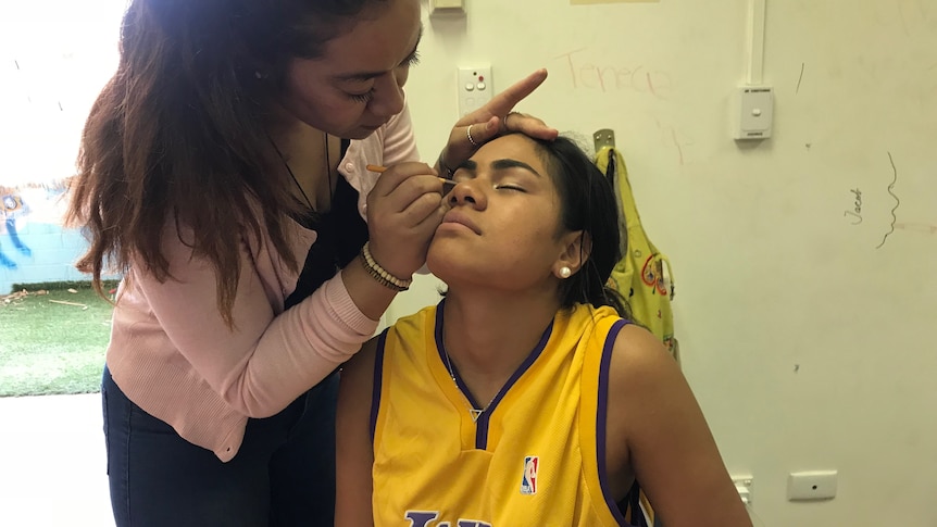 A woman leans over to apply eye make up to a girl who is sitting in a chair with her eyes closed.