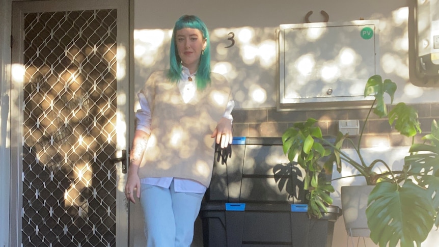 Madi  - a young woman with straight green-blue hair leaning against a large black storage crate on a house porch