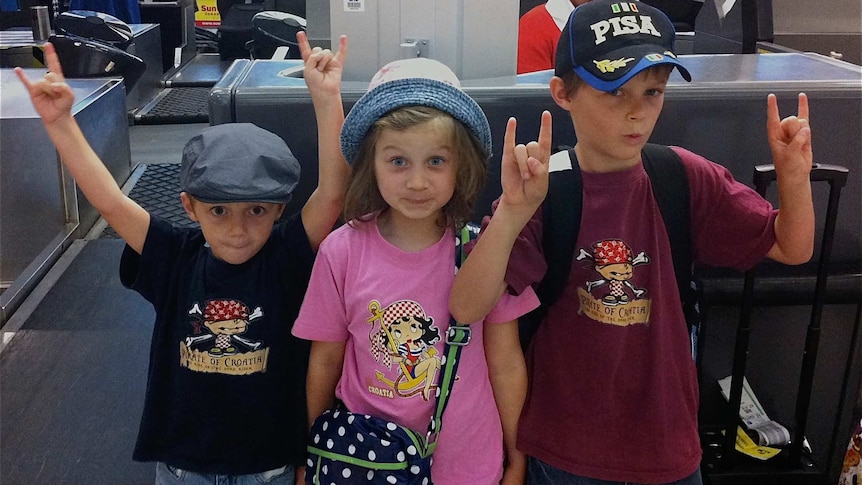Three children, all wearing hats, stand next to an airport check in counter