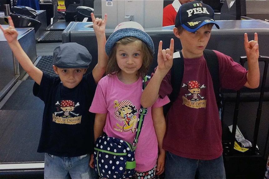 Three children, all wearing hats, stand next to an airport check in counter