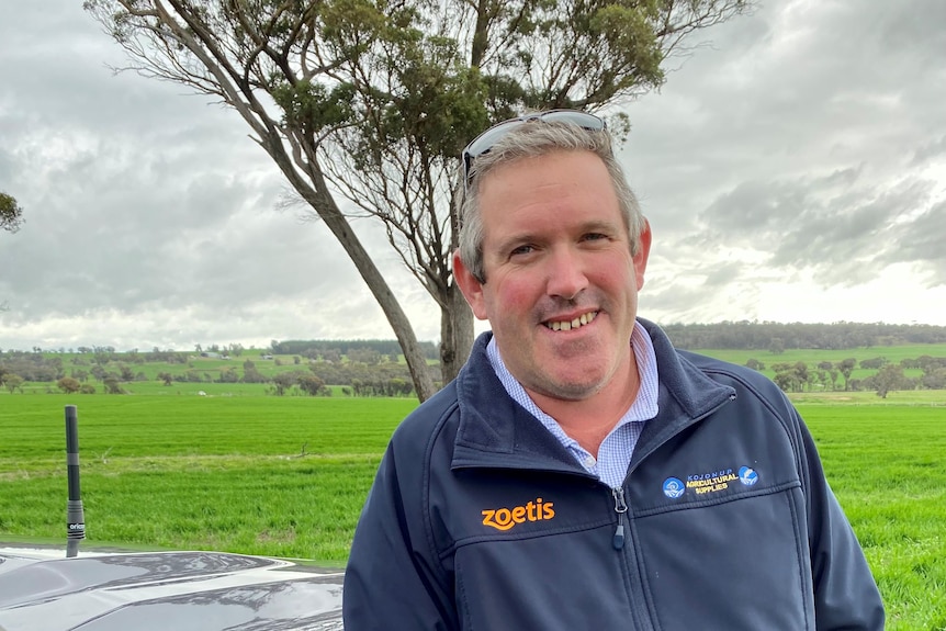 A middle-aged man smiles while leaning against a ute with a tree behind