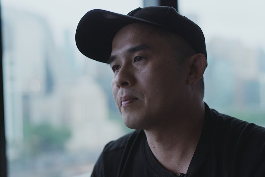 A man in a black baseball cap and black T-shirt sits in front of a window.