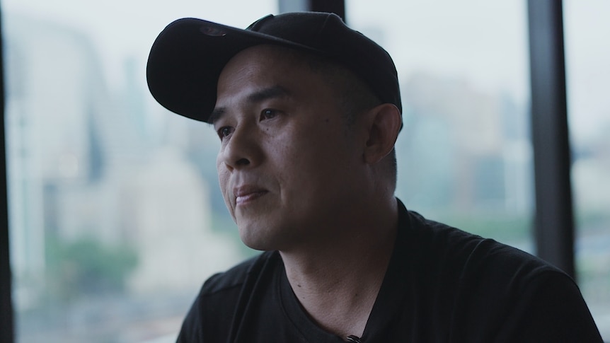 A man in a black baseball cap and black T-shirt sits in front of a window.