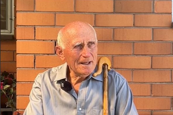 An elderly man sits in front of a brick wall with a walking stick resting on his shoulder.