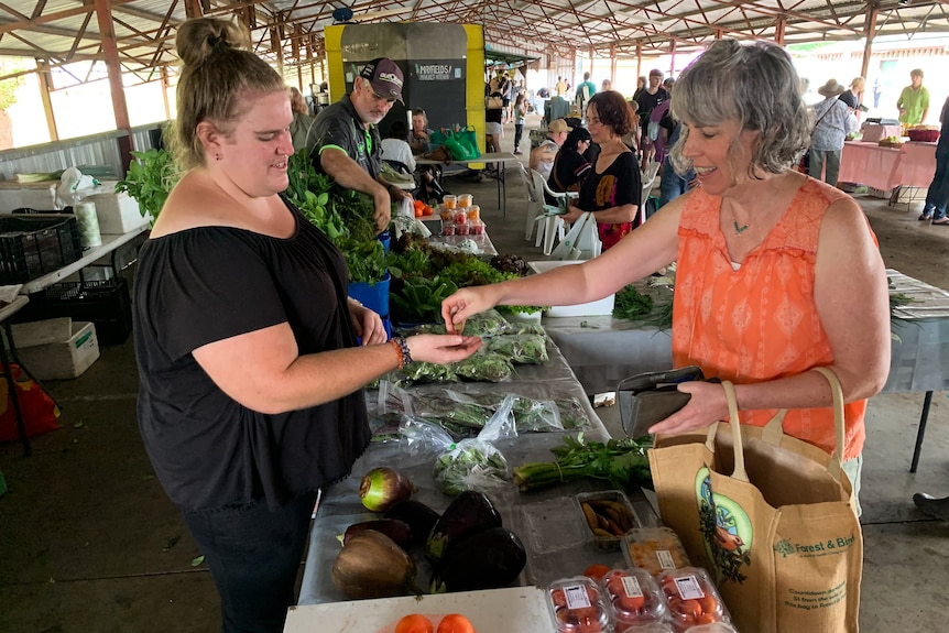 A woman in a black shirt sells vegetables to a woman in an orange shirt.