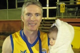 A man in a yellow football guernsey holds a baby and a beer, with a medal around his neck.