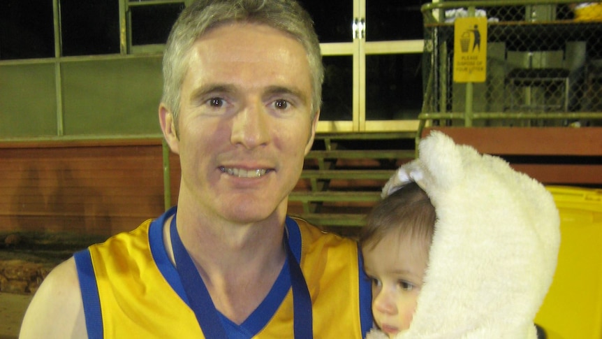 A man in a yellow football guernsey holds a baby and a beer, with a medal around his neck.