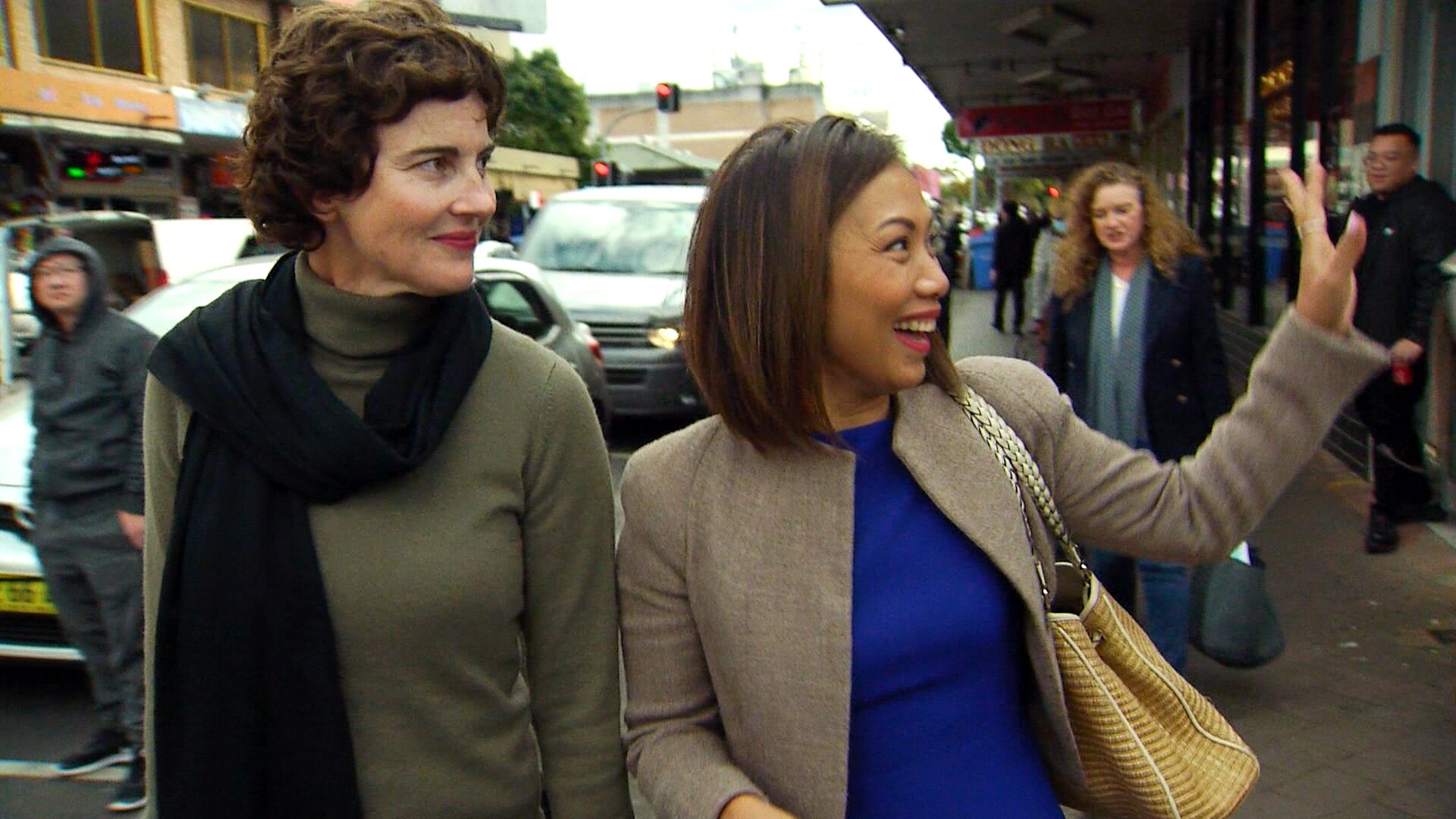 Kate Chaney in a green turtle neck and black scarf smiles and Dai Le smiles and waves as they walk along footpath