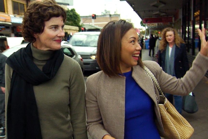 Kate Chaney in a green turtle neck and black scarf smiles and Dai Le smiles and waves as they walk along footpath