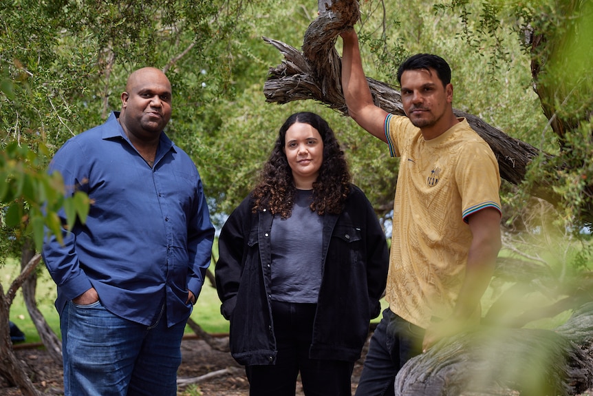 Three First Nations people, two men on either side of a woman, stand near trees. They are looking into the camera lens.