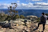 A bushwalker stands with their foot on a rock looking at a vast view, clouds in the distance.