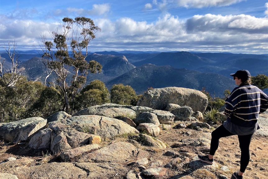 A bushwalker stands with their foot on a rock looking at a vast view, clouds in the distance.
