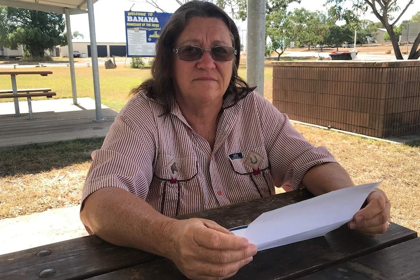 A woman sits a picnic table looking at the camera while holding a letter