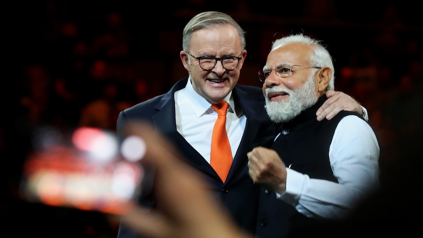 Anthony Albanese smiles as he puts his arm around Narendra Modi. Modi is making a fist. The background behind them is dark.