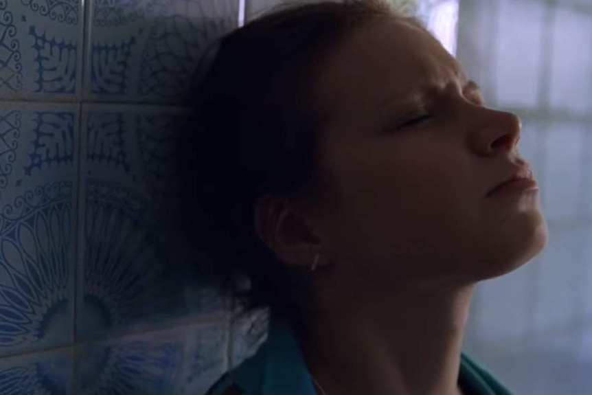 A woman leans her head against a tiled wall with a pained expression on her face.