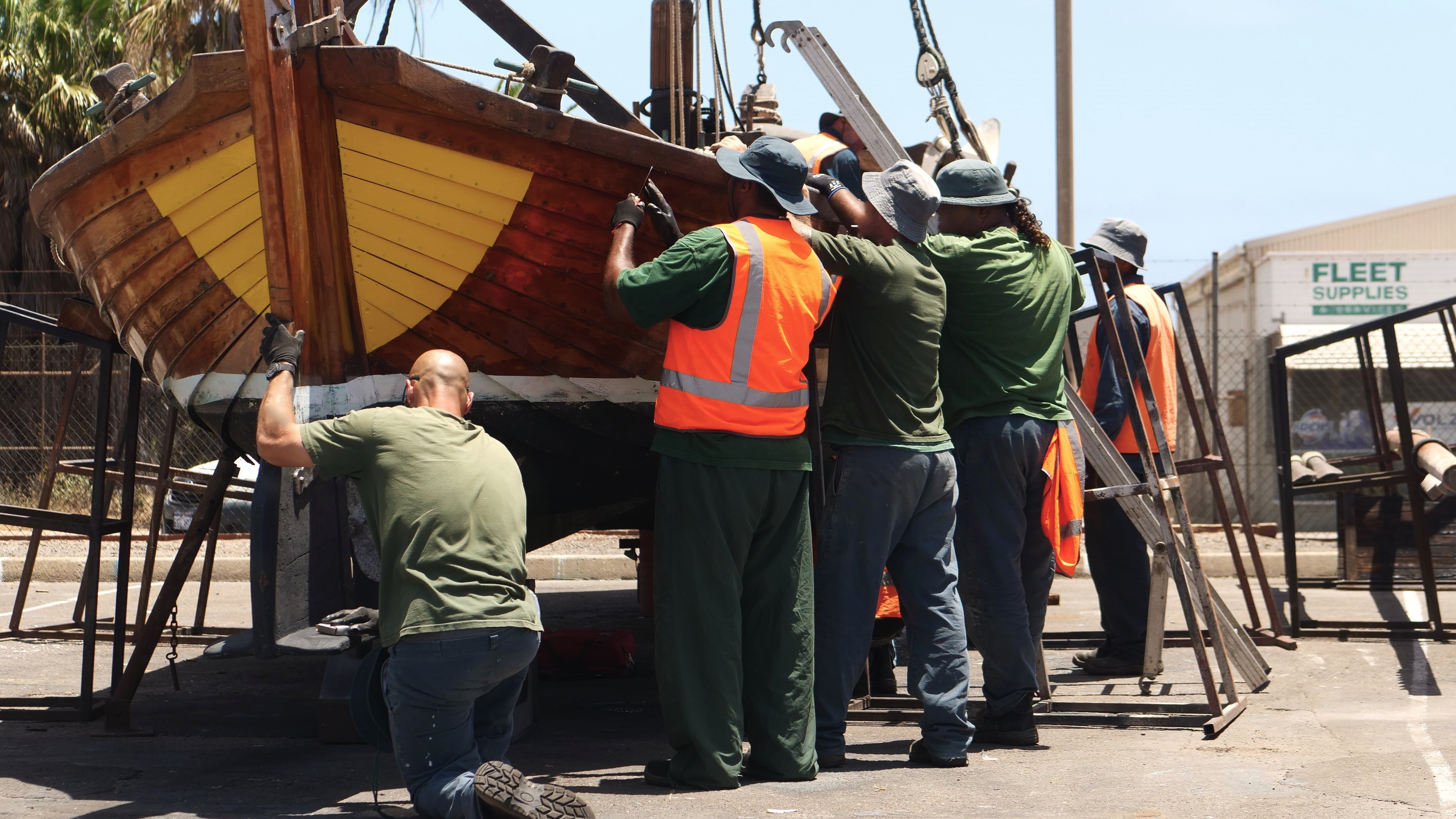 A group of men with backs to the camera working on the side of a wooden vessel on a hardstanc