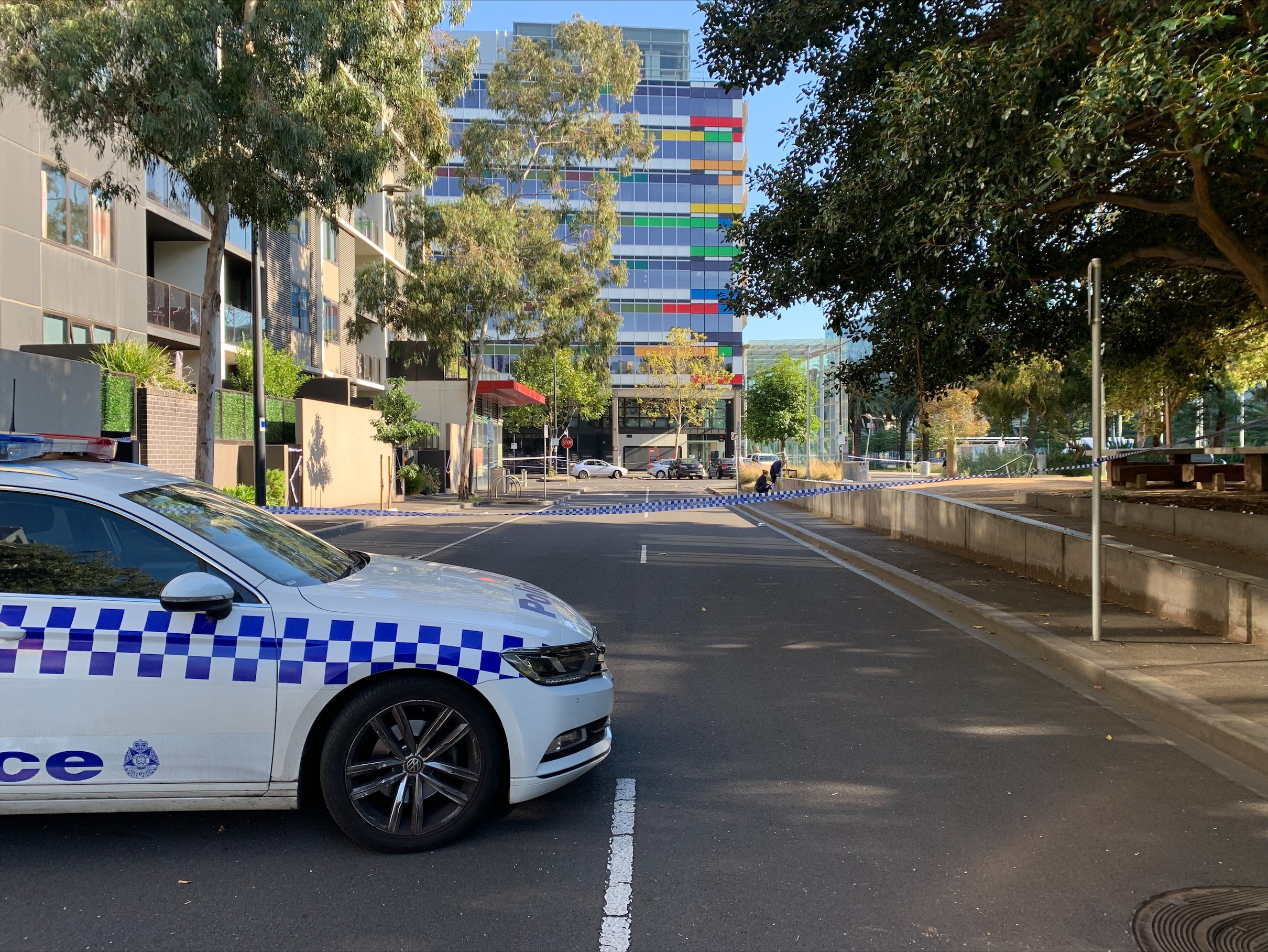 A police car is parked on a cordoned off street. Trees line one side of the road and a high-rise building is at the end.