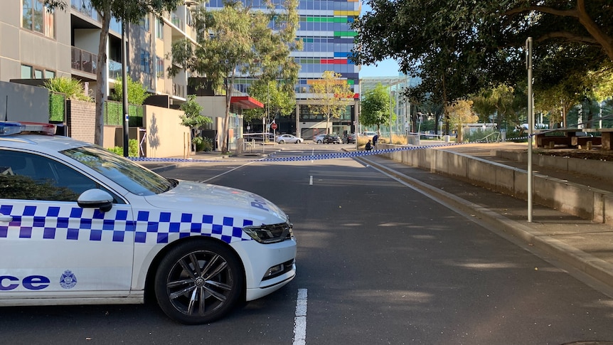 A police car is parked on a cordoned off street. Trees line one side of the road and a high-rise building is at the end.
