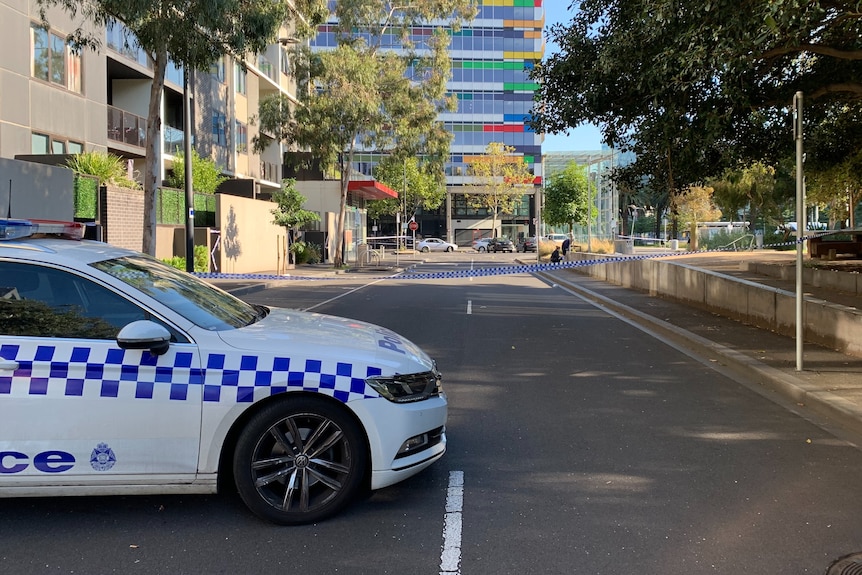 A police car is parked on a cordoned off street. Trees line one side of the road and a high-rise building is at the end.