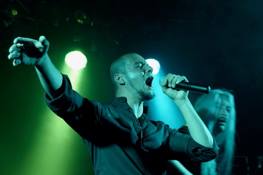 A bald man singing passionately into a microphone on a stage basked in green and blue light.