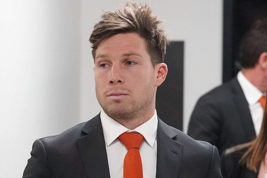A male AFL player wearing a dark suit, white shirt and orange tie, walks with his hands in his pockets.