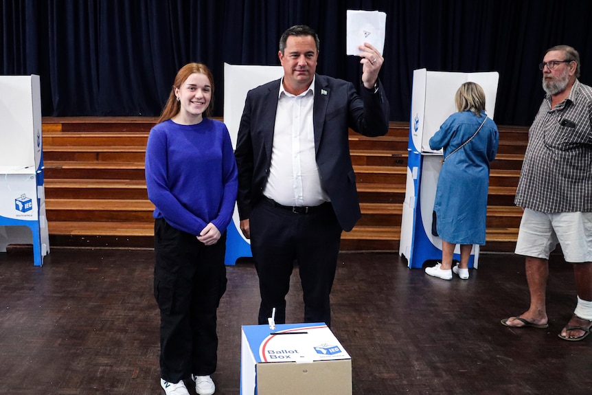 A man in a suit stands smiling, holding a paper ballot in front of a cardboard box with a young woman standing next to him 