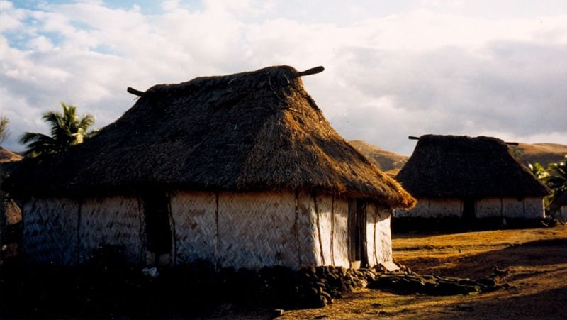 A hut is seen in the foreground of the image with another to the right, behind. The sky overhead is blue and cloudy.