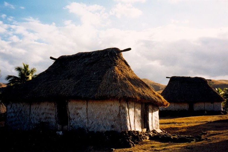A hut is seen in the foreground of the image with another to the right, behind. The sky overhead is blue and cloudy.
