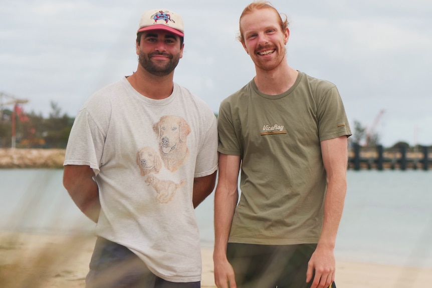 Two men smile at the camera with beach in the background. 