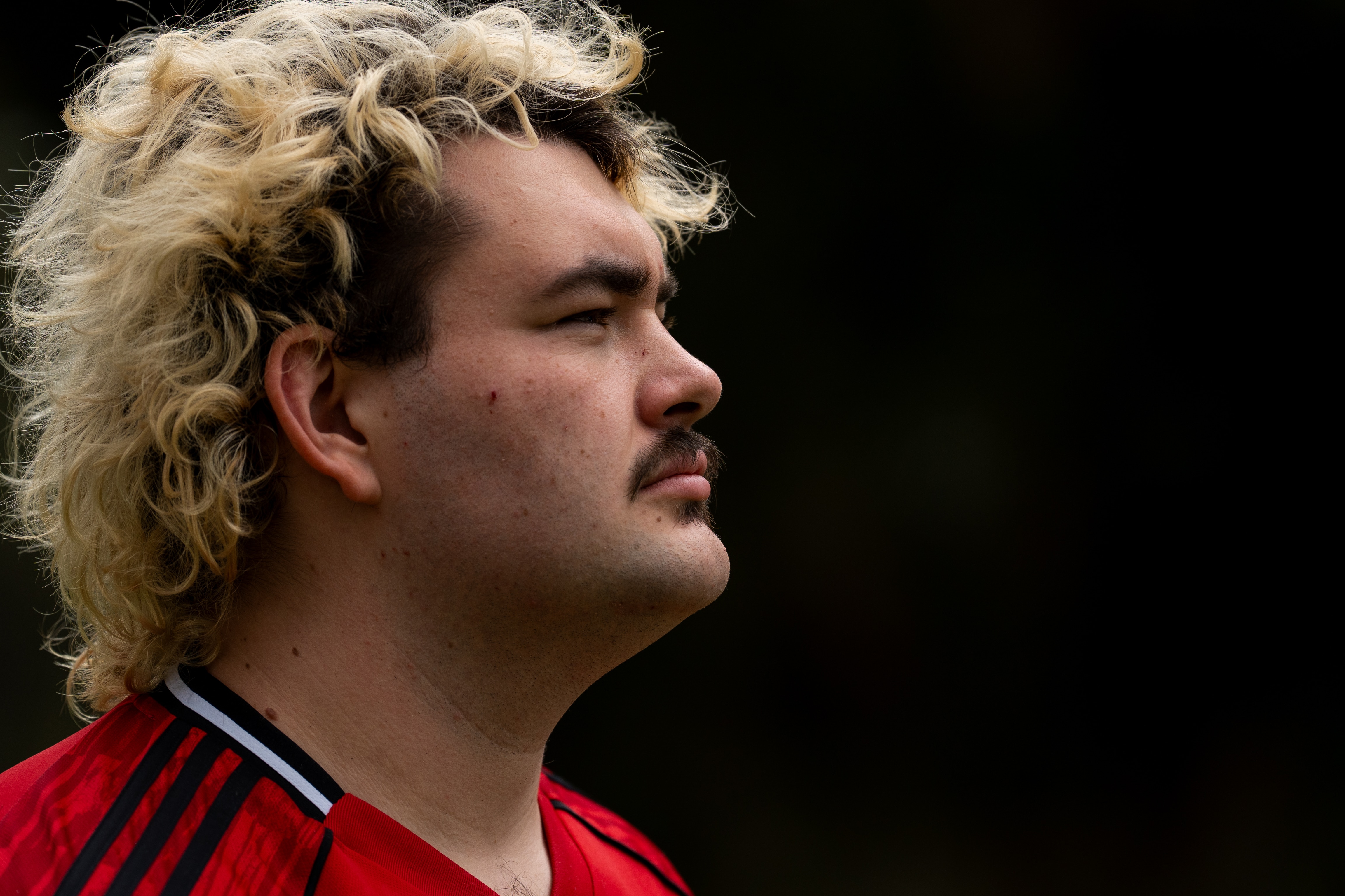 Um homem com cabelos loiros em um fundo escuro olhando para longe.