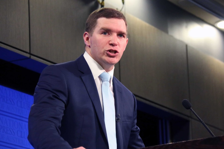 A suited man stands at a podium speaking.