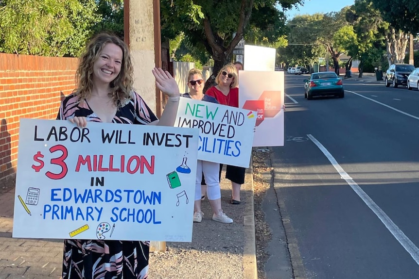 A row of women standing on the side of the road holding signs promising funding for a school