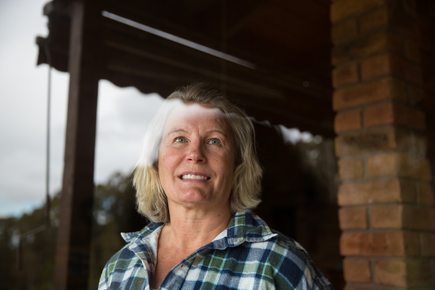 A woman looks out through a window, bush reflected in the glass.