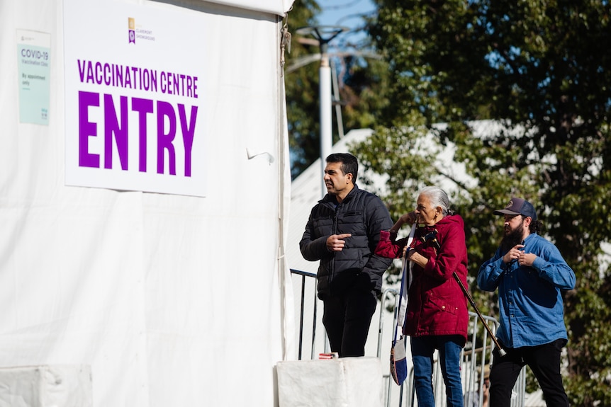 A woman is flanked by two men in a queue outside the entry to a vaccination centre at a pavilion at Claremont Showground.