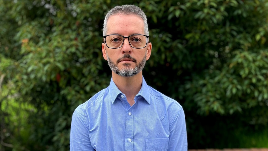 A bespectacled man with short grey hair and a neat beard stands outdoors, looking solemn.