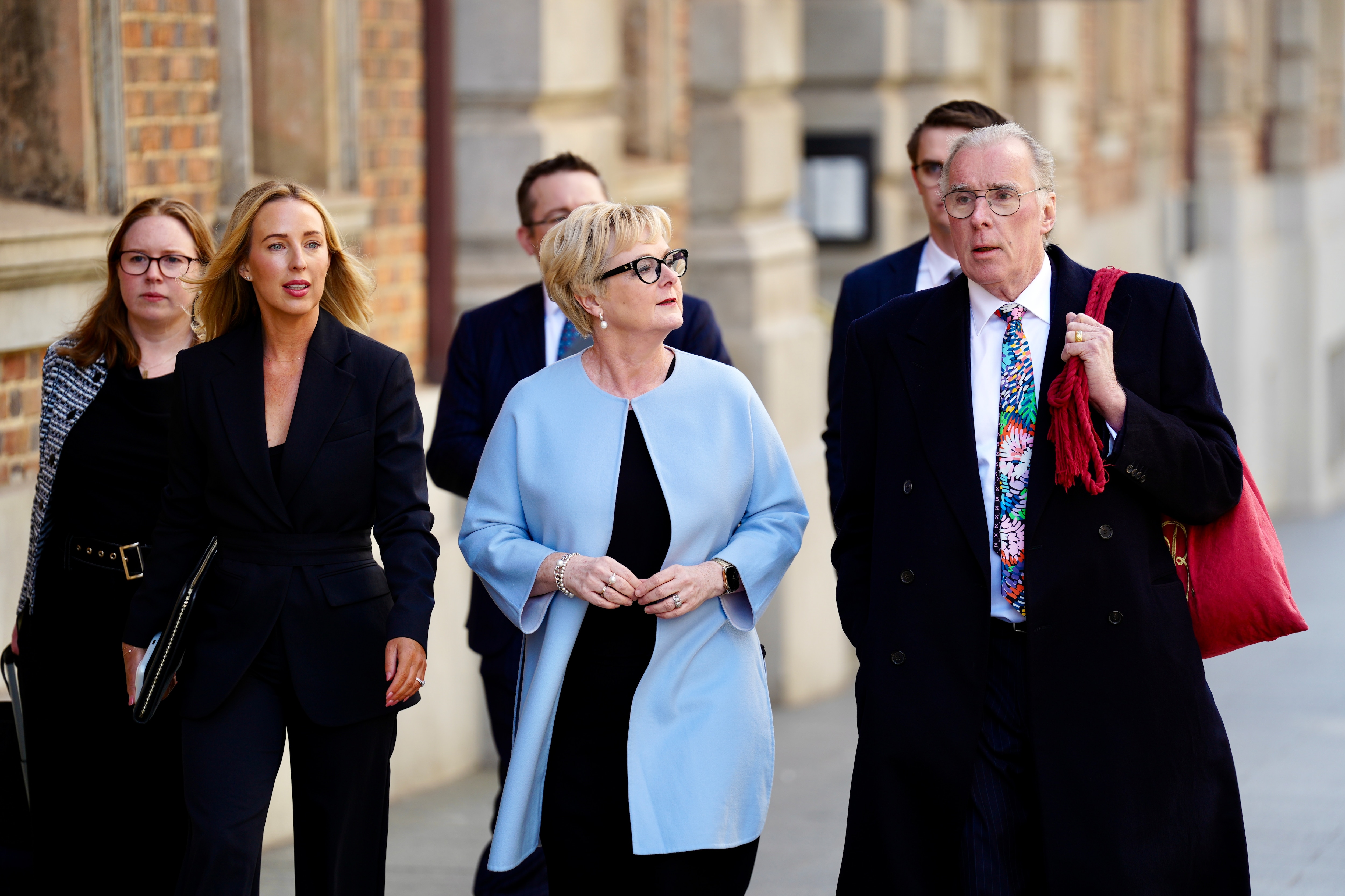 Linda Reynolds chega ao tribunal entre um grupo de pessoas, vestida com terno azul e preto e óculos.