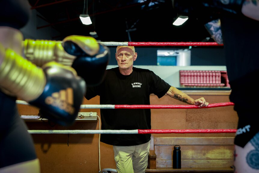 Boxing coach Kel Bryant, standing on the side of a ring in a gym staring at the camera