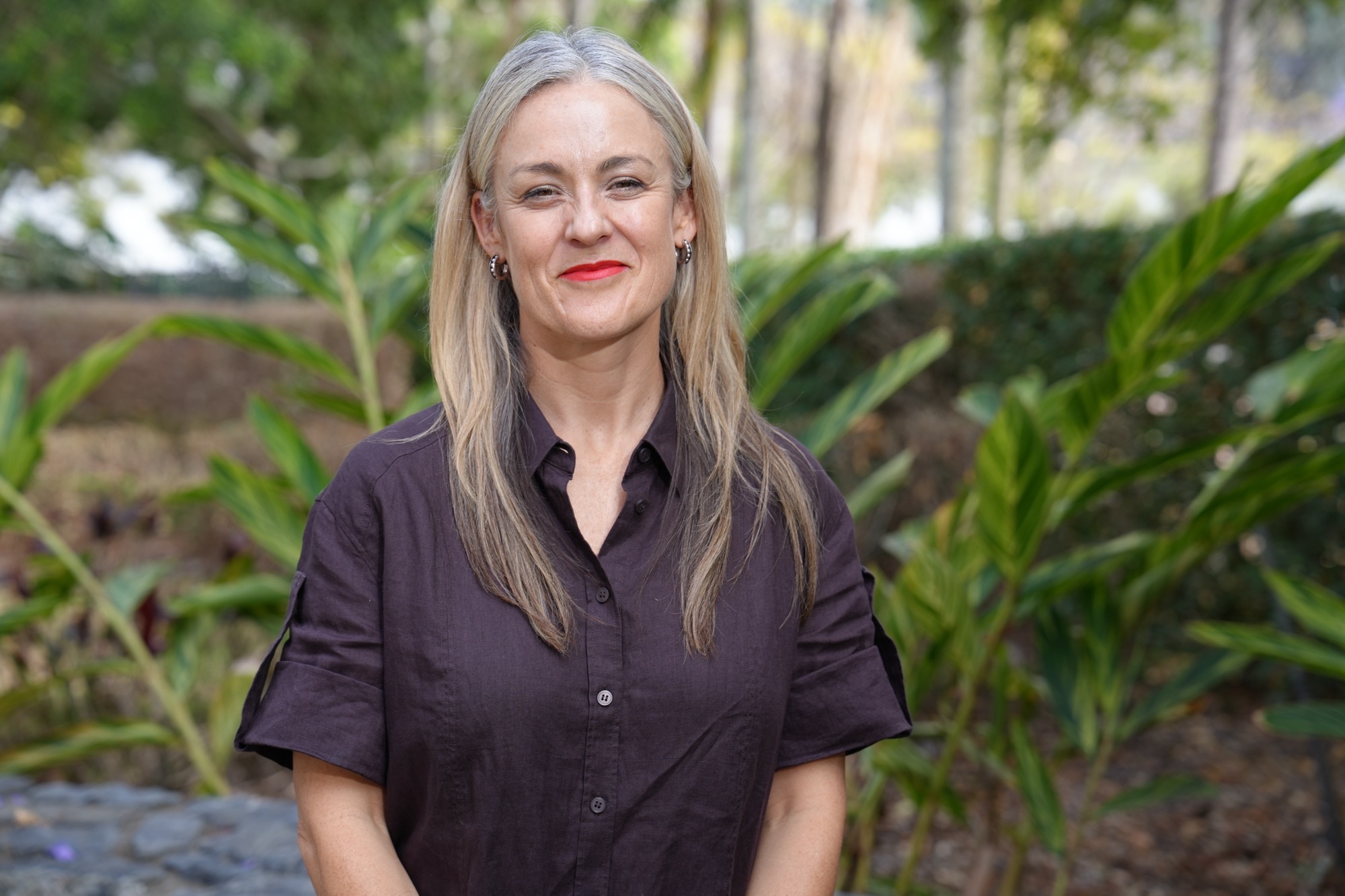 A portrait of a woman with blonde hairing wearing a brown blouse with trees and greenery in the background.