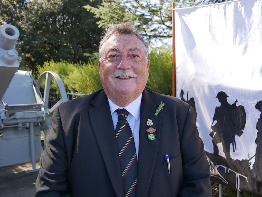 An older man stands outside an anzac day memorial wearing a suit.