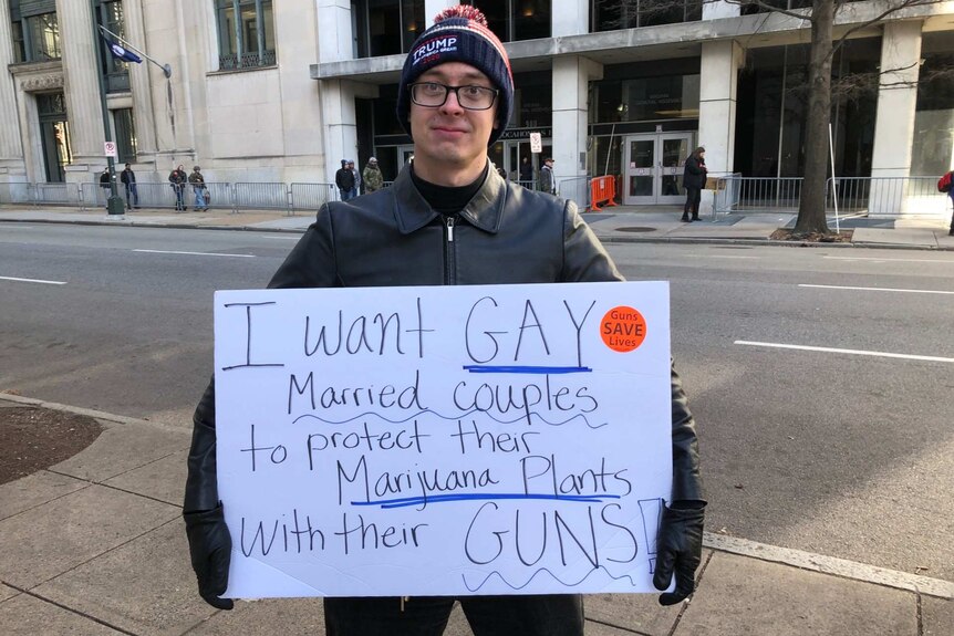 a man wearing a Trump beanie holds a sign that says i want gay married couples to protect their marijuana plants with their guns