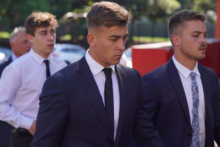 A young man in a suit, flanked by two others of similar age, walking.