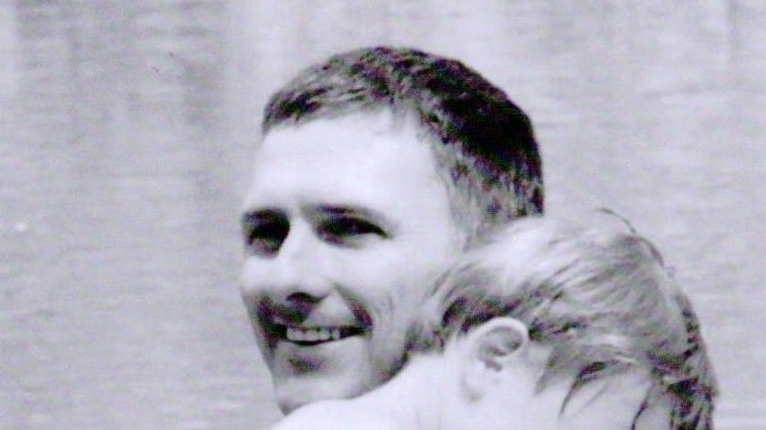 A black and white photo of Karm smiling with a toddler-aged child over his shoulder.