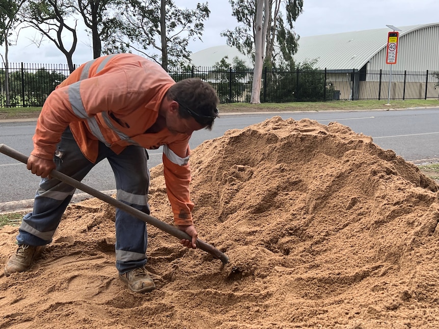 A man in orange hi-vis digs from a pile of sand. 