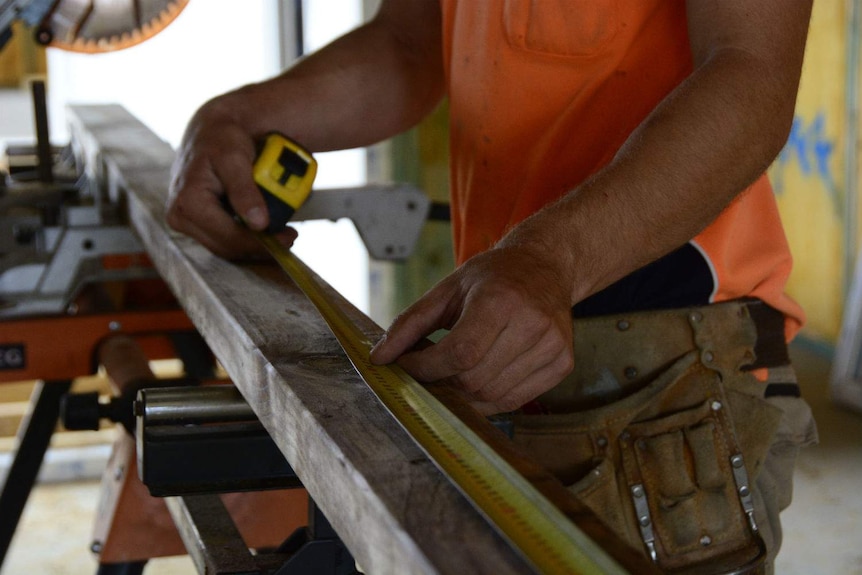 A close up of a man using a tape measure to measure a long piece of timber.