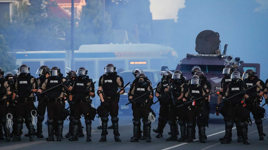 A row of heavily armed police stand in front of an armoured vehicle covered in smoke.